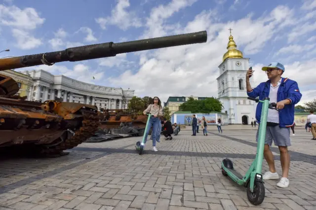 A man with a scooter takes a photo of a burnt-out tank