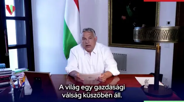 Orban speaking at a desk in an office with the Hungarian flag in the background