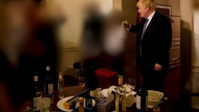 Boris Johnson raises a glass of drink to other people who are blurred out. In the foreground, there are bottles of alcohol littered on a table