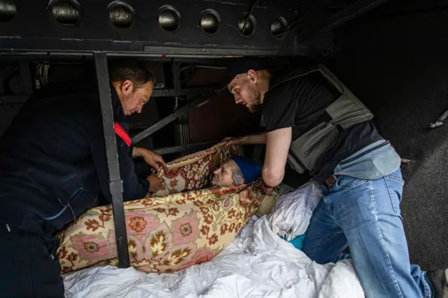 Two volunteers place an elderly woman who can't walk in the luggage space of an evacuation bus in Bakhmut.