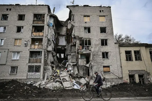 A man rides a bicycle past a destroyed residential block