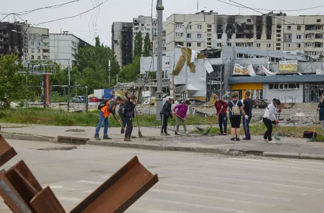 People in Kharkiv sweep a street with a damaged building behind