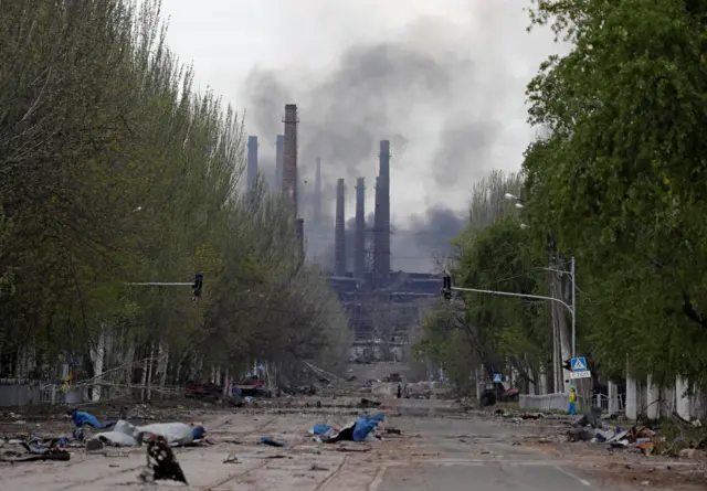 Debris litters a road leading to Azovstal, with smoke rising from the complex in the background