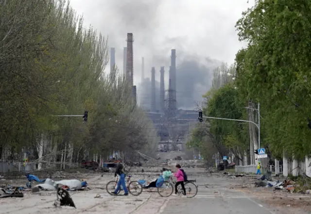 People walk their bikes across the street as smoke rises above a plant of Azovstal Iron and Steel Works in Mariupol on 2 May