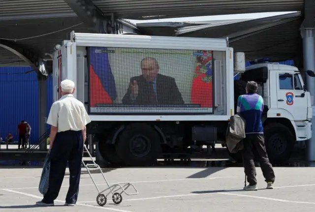 Russian President Vladimir Putin is seen on a big screen broadcasting Russian TV at a humanitarian aid distribution point