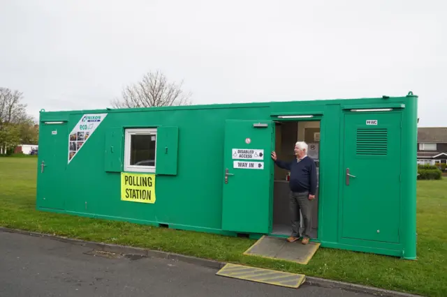 A man stands outside a green portable polling station