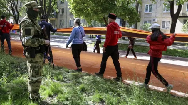 A man in military fatigues holding a gun looks on as people carry a St George's ribbon through Mariupol