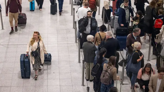 A woman walks with a suitcase at an airport next to a queue of people