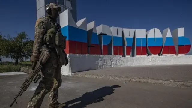 A picture taken during a visit to Mariupol organised by the Russian military shows a Russian serviceman walks in front of the re-painted in colours of Russian National flag sign "Mariupol" at the entrance to the city of Mariupol, Donetsk region