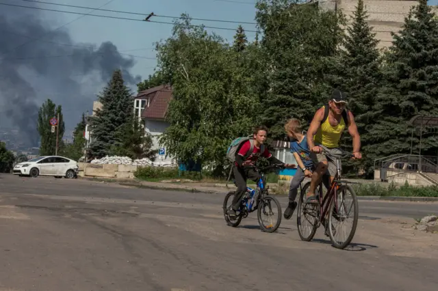 A man and two boys cycle along an empty street, as smoke rises after shelling in the background, Lysychansk, 10 June 2022