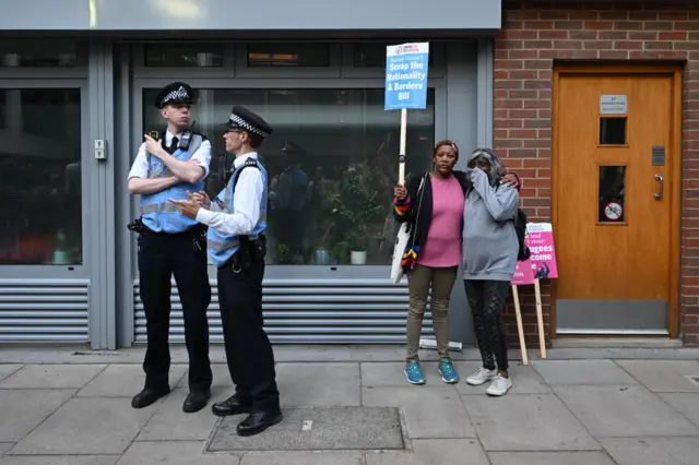 A woman holds a placard saying 'Scrap the nationalities and borders bill' as they are seen behind police officers during a protest against the UK deportation flights to Rwanda outside the Home Office