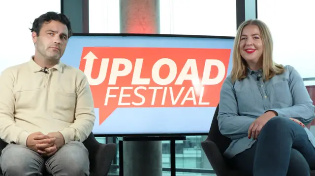 male and female presenters sit infront of a sign that reads "Upload Festival".