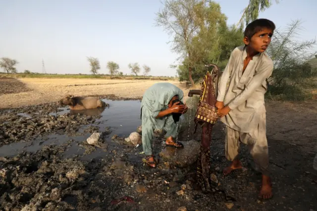 One boy bathes while another draws water from a hand pump, near a buffalo sitting in the mud to cool off, during a heatwave, in Jacobabad, Pakistan