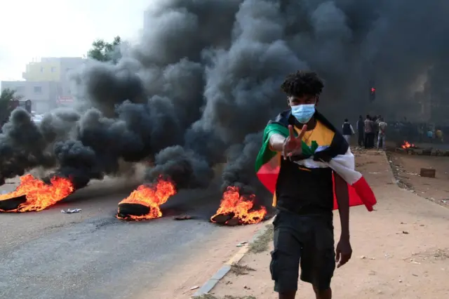 A Sudanese protester draped with the national flag flashes the victory sign next to burning tyres during a demonstration in the capital Khartoum, on October 25, 2021