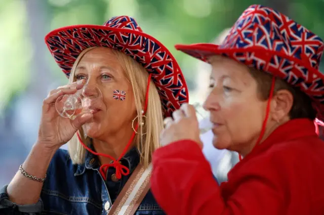 Women wearing Union Jack hats, and drinking bubbly beverages, get ready along The Mall