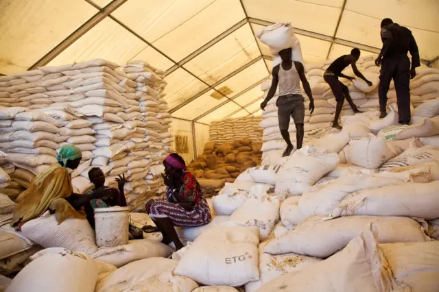 Men carry bags of food while women wait for their rations at the WFP food distribution site in Pibor