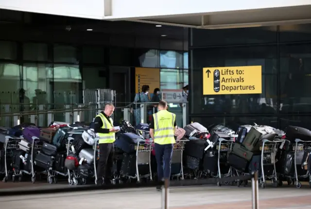 Airport workers stand next to lines of passenger luggage arranged outside Terminal 2 at Heathrow Airport in London