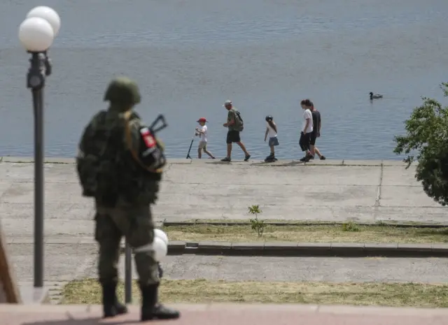 A Russian serviceman standing guard as a family walks on a promenade along the Dnipro River in Kherson