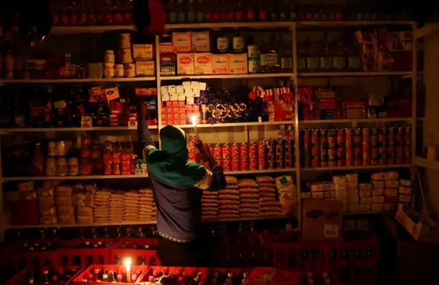 A shop owner picks an item for a customer by candlelight during power outages