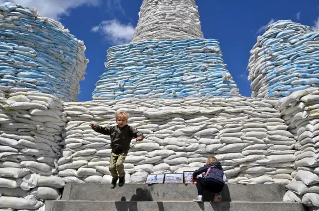 Children jumping and playing around a wall of sandbags