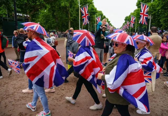 A group of spectators wearing Union Flag hats and capes cross the Mall