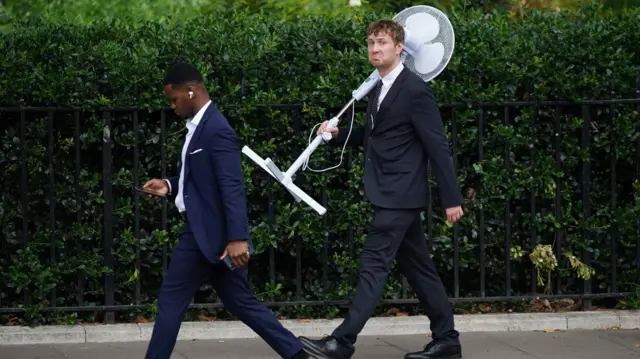 Two men walking in suits. One holds a large fan and has a sad face.