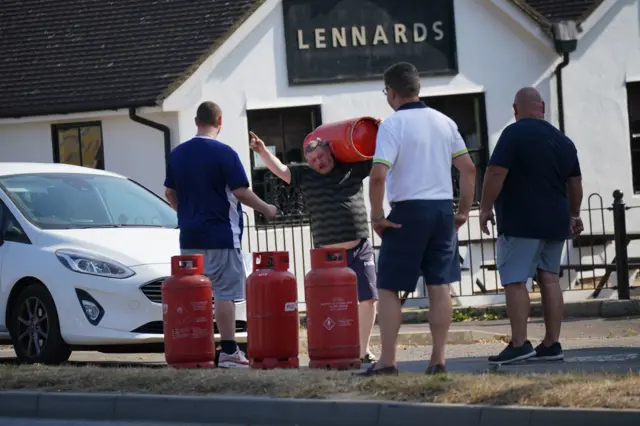 Three men stand around large red gas canisters as a man carries one over his shoulder and gestures to the group