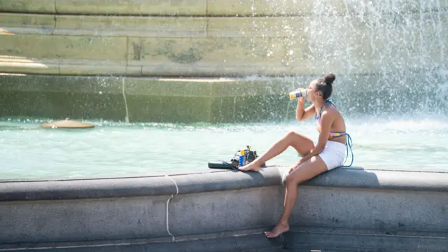 A woman drinks water as she sits in the spray of a fountain in Trafalgar Square