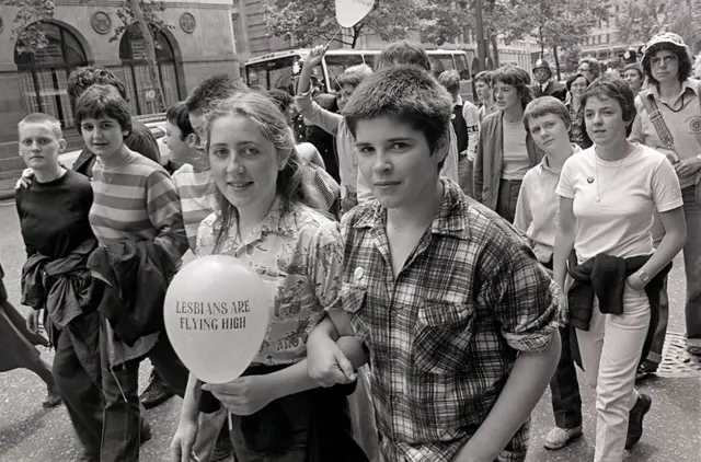 A crowd of people attend a Pride event in 1980, with one person holding a balloon that says 'lesbians are flying high'