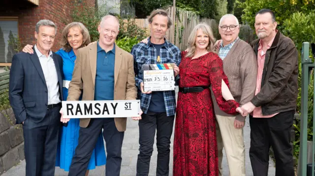 Neighbours 80s alumni stand with a sign of Ramsay Street and a clapperboard