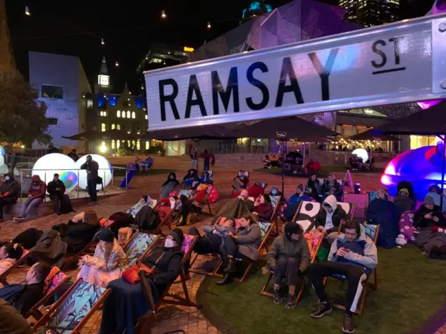 People dressed up in warm clothes sit in deck chairs to watch the finale, with a Ramsay Street sign in the foreground