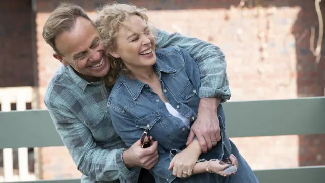 In character, Donovan hugs Minogue from behind as they both smile