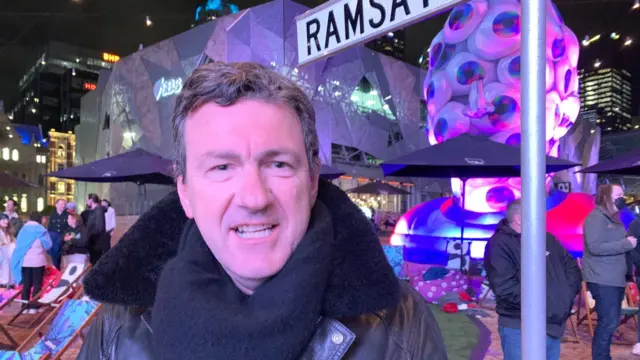 Iain Batty in a coat and a scarf, stood next to the Ramsay Street sign at the screening of the Neighbours final in Melbourne