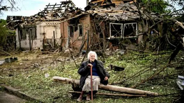 A woman sits in front of a destroyed house