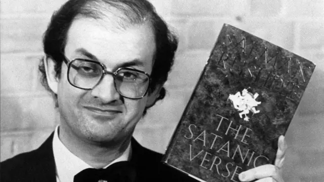 Salman Rushdie holds up a copy of his book, The Satanic Verses, while wearing a suit, in 1989