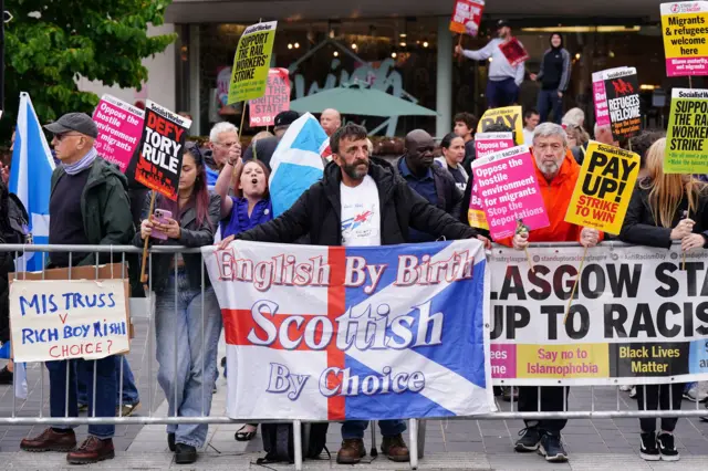 Protestors gather outside the venue - front and centre is a man holding a flag which says "English by birth, Scottish by choice"