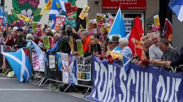 A group of protesters with placards and flags gather behind a security barrier