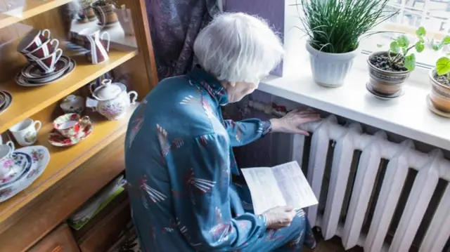 An elderly woman holding a gas bill in front of heating radiator