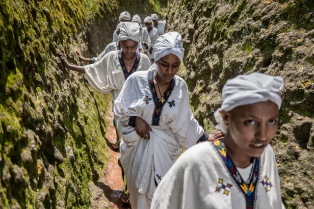 Young women in white robes walk through a narrow path between stone walls.