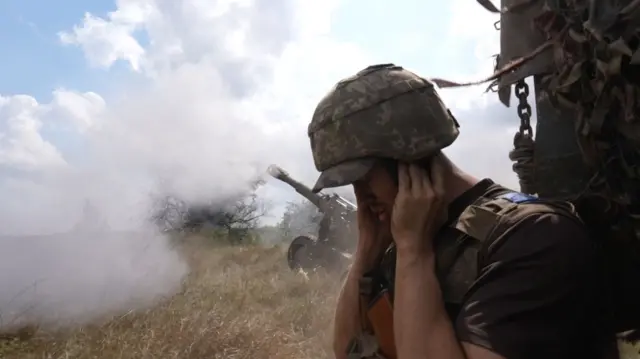 A soldier covers his ears as a howitzer goes off behind him