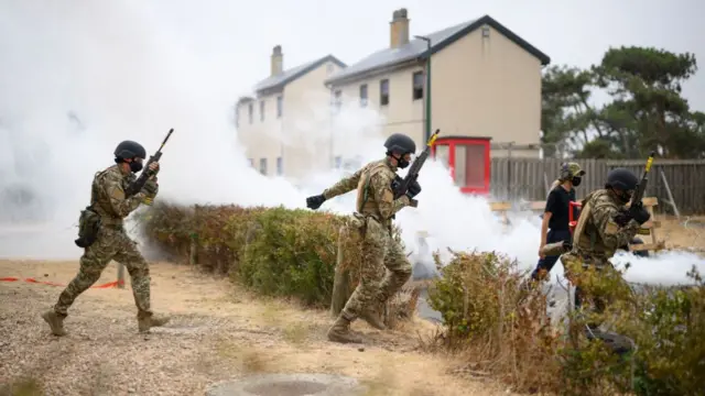 Three soldiers in masks and helmets run across a smokey patch of land