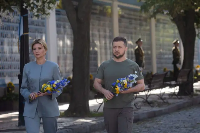 Zelensky and wife look solemn as they carry blue and yellow flowers