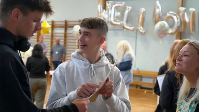 A student receiving his results last year, looking pleased, with a Well Done banner in the background