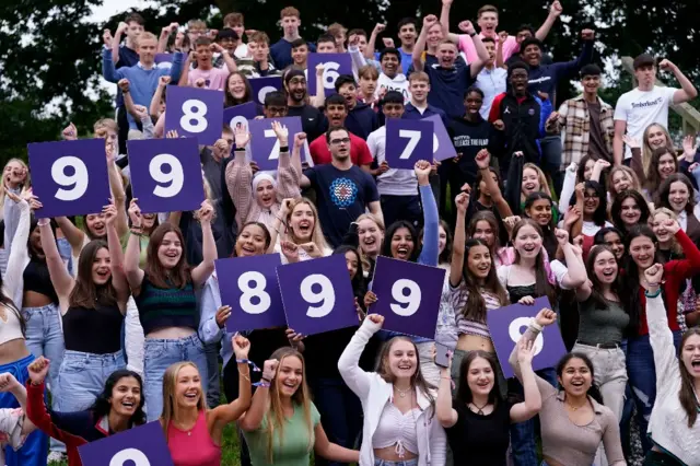 A large group of students hold their fists in the air and hold aloft boards showing numbers, including 7, 8 and 9