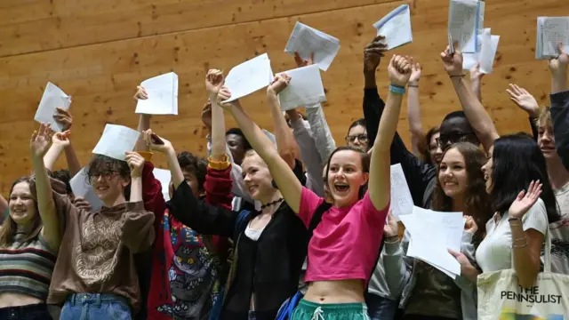 A large group of teenagers hold papers with their exam grades in the air