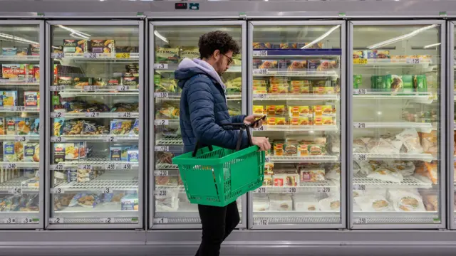 A man, holding a green shopping basket, stands in front of a row at freezers at a supermarket