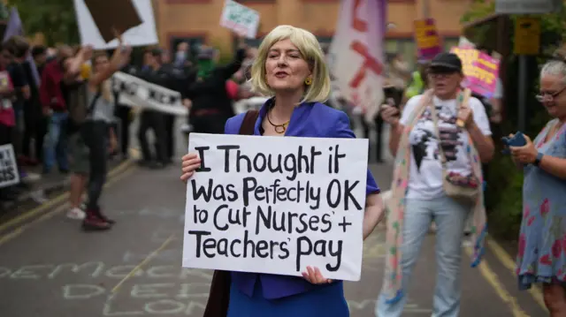 A woman dressed as Liz Truss - in a blonde wig and a blue suit - holds a placard saying "I thought it was perfectly OK to cut nurses' and teachers' pay"