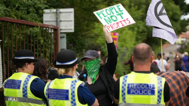 A demonstrator holds a sign saying "people not profits" in front of three police officers