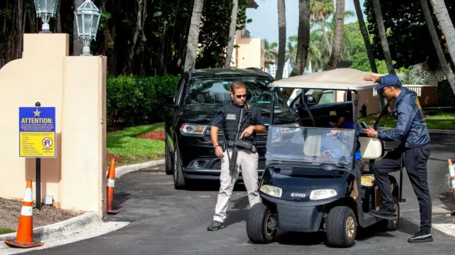 A secret service guard with a rifle is seen standing next to two other guards and a golf buggy