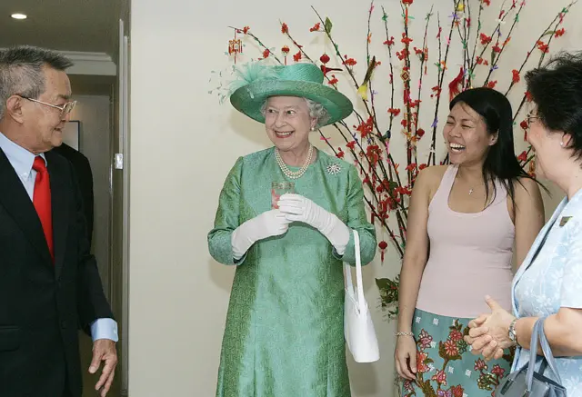 Britain's Queen Elizabeth (C) holds a glass of water as she poses with the Pung family, Thomas (L) and his daughter Genevieve (R) during a visit to their home in Singapore, 17 March 2006, the same glass was presented to her by the family during The Queen's 1972 visit to their housing project.